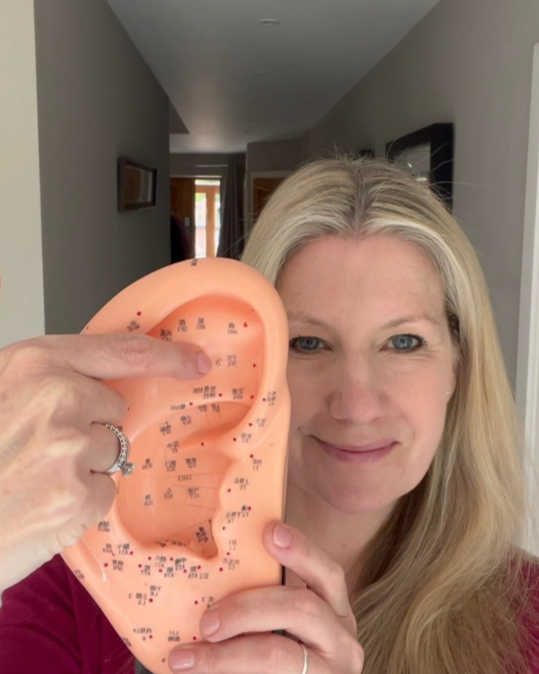 A woman with blonde hair holds up a peach-colored silicone ear model showing acupuncture or massage points, with her finger pointing to details on the ear. The background shows a home hallway.